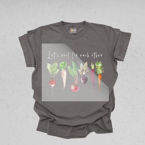 K&ouml;nnte beinhalten: Ein dunkelgraues T-Shirt mit einer Aquarell-Illustration von Gem&uuml;se und dem Text "Let's root for each other".