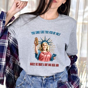 May include: A heather gray t-shirt featuring a graphic of a woman dressed as the Statue of Liberty holding a hot dog. The text on the shirt reads "YOU LOOK LIKE THE 4TH OF JULY MAKES HE WANT A HOT DOG REAL BAD."