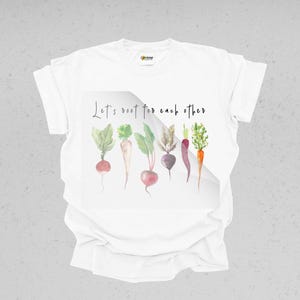 K&ouml;nnte beinhalten: Wei&szlig;es T-Shirt mit einer Aquarell-Illustration von Gem&uuml;se und dem Text "Let's root for each other".