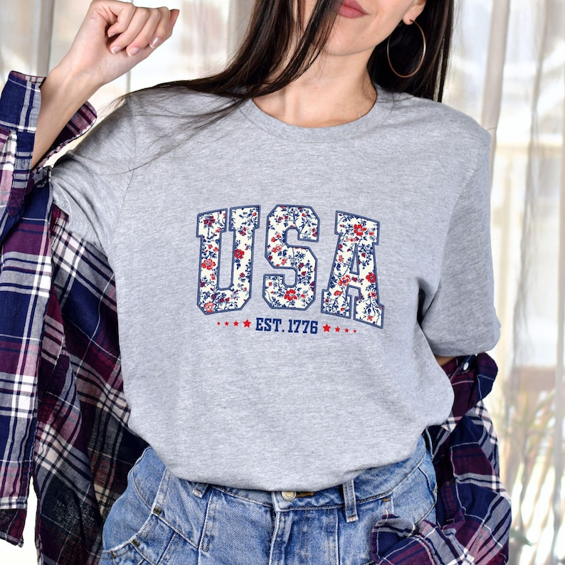 May include: A heather gray t-shirt featuring the word "USA" in a floral pattern with red and blue accents. Below the word "USA" is the text "EST. 1776" with stars. The shirt is a crew neck style.