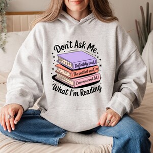 May include: A light gray hoodie with the text "Don't Ask Me What I'm Reading" and a stack of three books. The books are purple, peach, and pink, with the titles "Definitely smut," "The smittiest smut," and "Even more smut lol."