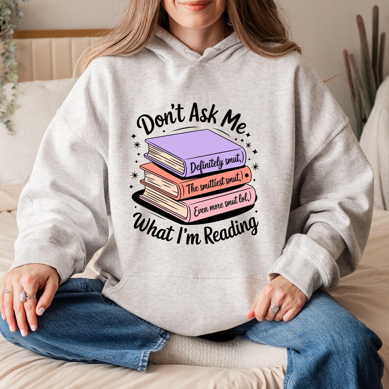 May include: A light gray hoodie with the text "Don't Ask Me What I'm Reading" and a stack of three books. The books are purple, peach, and pink, with the titles "Definitely smut," "The smittiest smut," and "Even more smut lol."