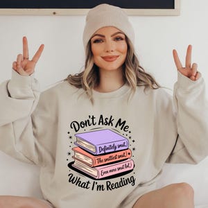 May include: Beige sweatshirt with the text "Don't Ask Me What I'm Reading" and a stack of books. The books are purple, peach, and white, with text that reads "Definitely smut," "The smuttiest smut," and "Even more smut lol." The person is wearing a beige beanie.