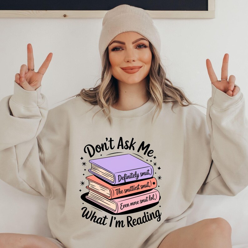 May include: Beige sweatshirt with the text "Don't Ask Me What I'm Reading" and a stack of books. The books are purple, peach, and white, with text that reads "Definitely smut," "The smuttiest smut," and "Even more smut lol." The person is wearing a beige beanie.