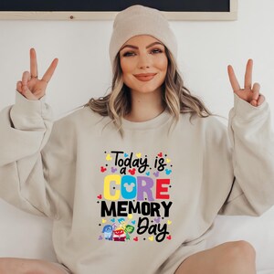 May include: A beige sweatshirt with the text "Today is a CORE MEMORY Day" in colorful letters. The text is surrounded by small Mickey Mouse heads and stars. There are five characters from the movie Inside Out on the bottom of the sweatshirt.