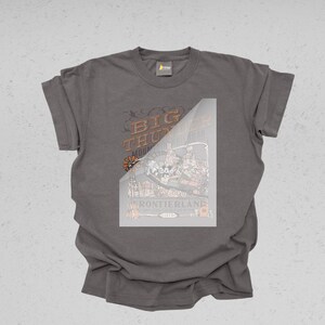 Peut inclure: Un t-shirt gris avec un motif graphique. Le motif pr&eacute;sente le texte "Big Thunder Mountain Railroad" et "Frontierland", ainsi que des illustrations d'un montagnes russes et d'autres &eacute;l&eacute;ments.