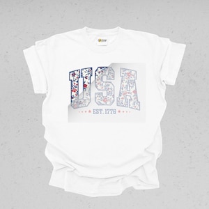 May include: White t-shirt featuring the word "USA" in a floral design. The letters are filled with a pattern of red, blue, and white flowers. Below the letters is the text "EST. 1776" with stars on each side.