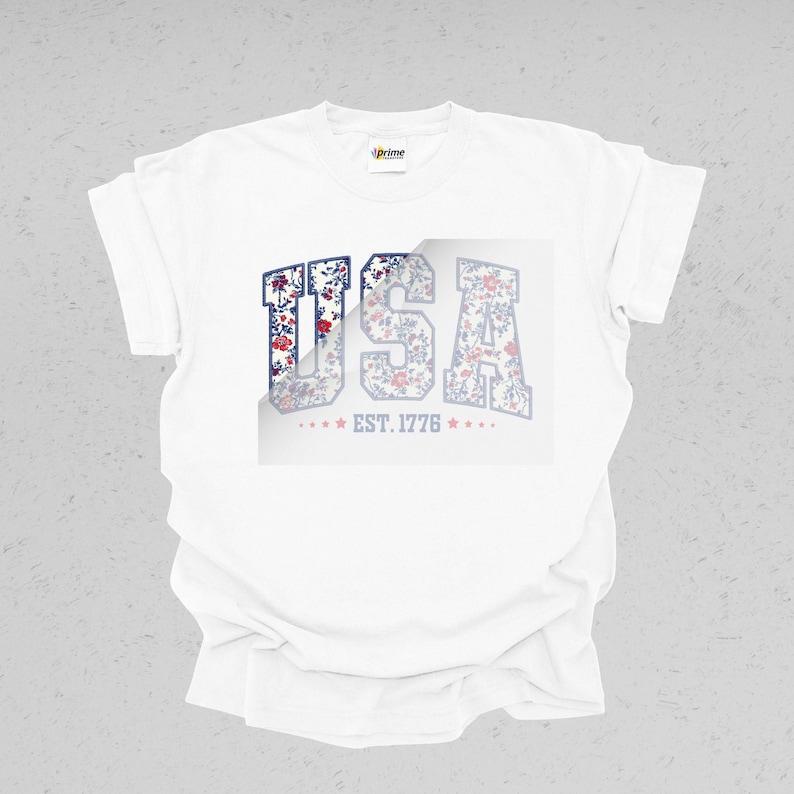 May include: White t-shirt featuring the word "USA" in a floral design. The letters are filled with a pattern of red, blue, and white flowers. Below the letters is the text "EST. 1776" with stars on each side.