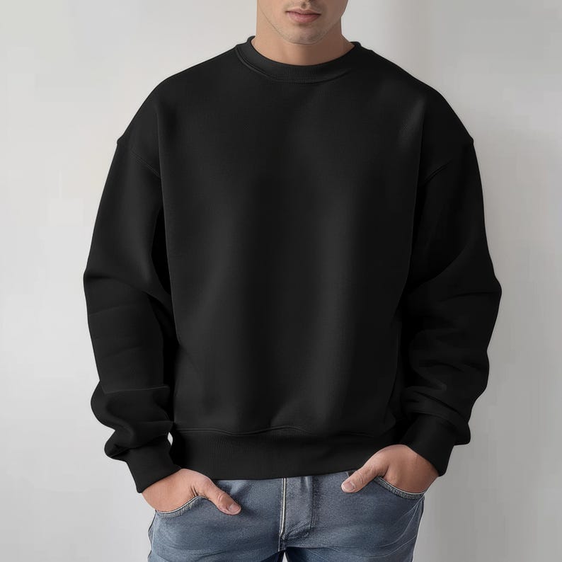 May include: A solid black crew neck sweatshirt. The sweatshirt has long sleeves and a ribbed hem and cuffs. The model is wearing blue jeans and has their hands in their pockets. The background is a plain white wall.