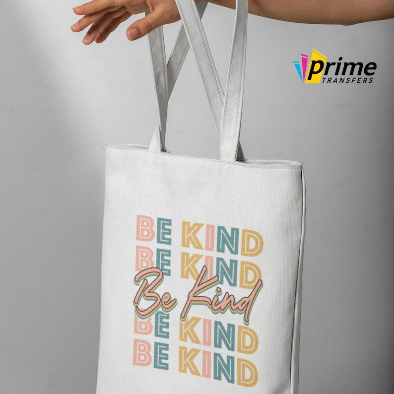 May include: White tote bag with the words "BE KIND" in pastel colors. The bag has two white handles and is held up by a hand. The bag is made of a canvas-like material.
