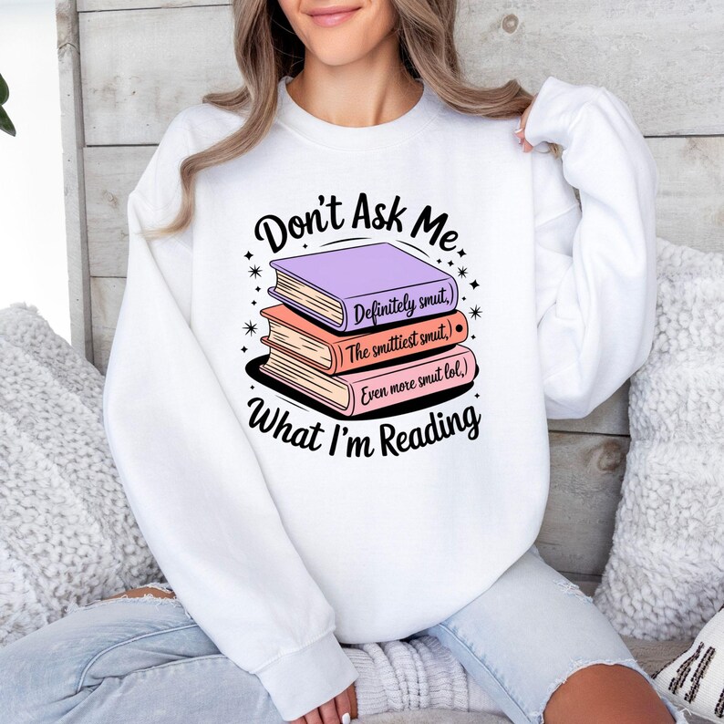May include: White sweatshirt with the text "Don't Ask Me What I'm Reading" and a stack of three books. The books are purple, peach, and pink, with the titles "Definitely smut," "The smuttiest smut," and "Even more smut lol."
