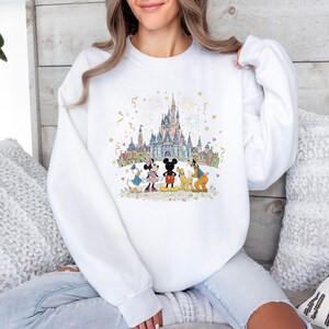 May include: White sweatshirt featuring a colorful illustration of a castle with fireworks and cartoon characters. The design includes Mickey and Minnie Mouse, Donald Duck, and Goofy. The sweatshirt has long sleeves and a crew neck.