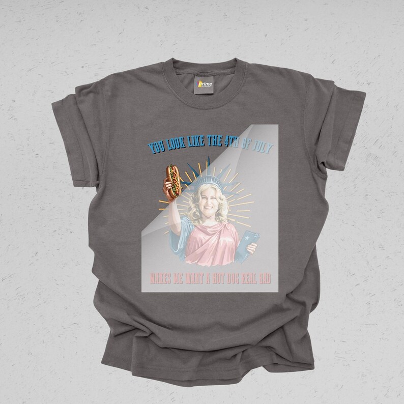 May include: Dark gray t-shirt with a graphic featuring a woman dressed as the Statue of Liberty holding a hot dog. Text reads "YOU LOOK LIKE THE 4TH OF JULY" and "MAKES ME WANT A HOT DOG REAL BAD."