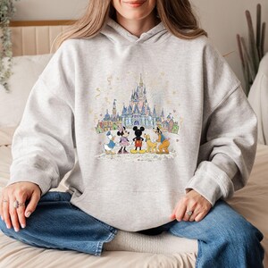 May include: A light gray hoodie featuring a colorful illustration of Disney characters in front of a castle. Mickey Mouse, Minnie Mouse, Donald Duck, Goofy, and Pluto are depicted. The design includes a castle, buildings, and confetti.