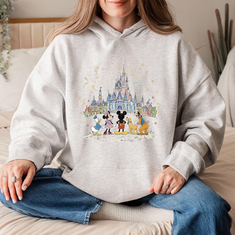 May include: A light gray hoodie featuring a colorful illustration of Disney characters in front of a castle. Mickey Mouse, Minnie Mouse, Donald Duck, Goofy, and Pluto are depicted. The design includes a castle, buildings, and confetti.