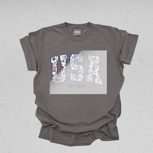 May include: A gray t-shirt with the word "USA" in a floral design. The letters are filled with a red, white, and blue floral pattern. Below the word "USA" is the text "EST. 1776" in a light red color.