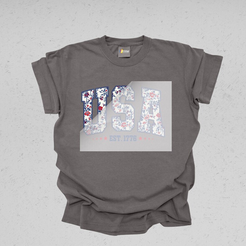 May include: A gray t-shirt with the word "USA" in a floral design. The letters are filled with a red, white, and blue floral pattern. Below the word "USA" is the text "EST. 1776" in a light red color.