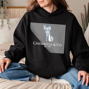 May include: Black hooded sweatshirt featuring a light blue high-heeled shoe and bow design on a gray square. The text "Cinderella & Co. Est 1950" is printed below the graphic. The sweatshirt is worn by a person.