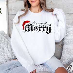 May include: White sweatshirt with the word "Merry" in black script, adorned with snowflakes and a Santa hat. The design is festive and suitable for the holiday season. The sweatshirt is a long-sleeved crew neck.