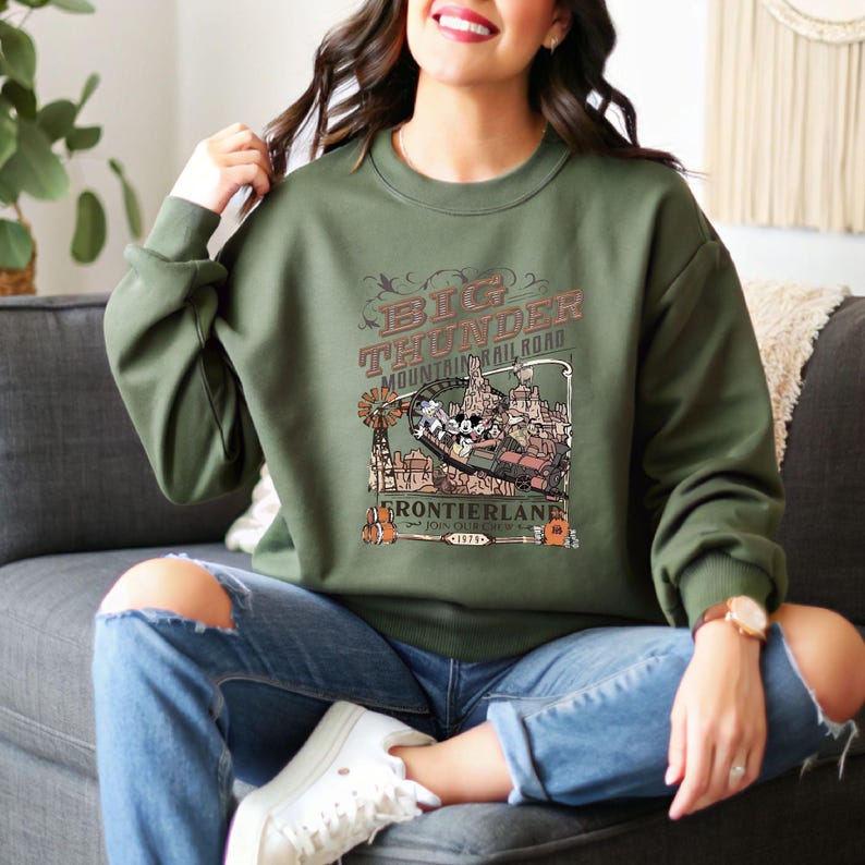 Peut inclure: Un sweat-shirt vert olive avec un motif graphique du man&egrave;ge Big Thunder Mountain Railroad. Le motif comprend le texte "Big Thunder Mountain Railroad" et "Frontierland" dans un style vintage. Le sweat-shirt est &agrave; col rond.