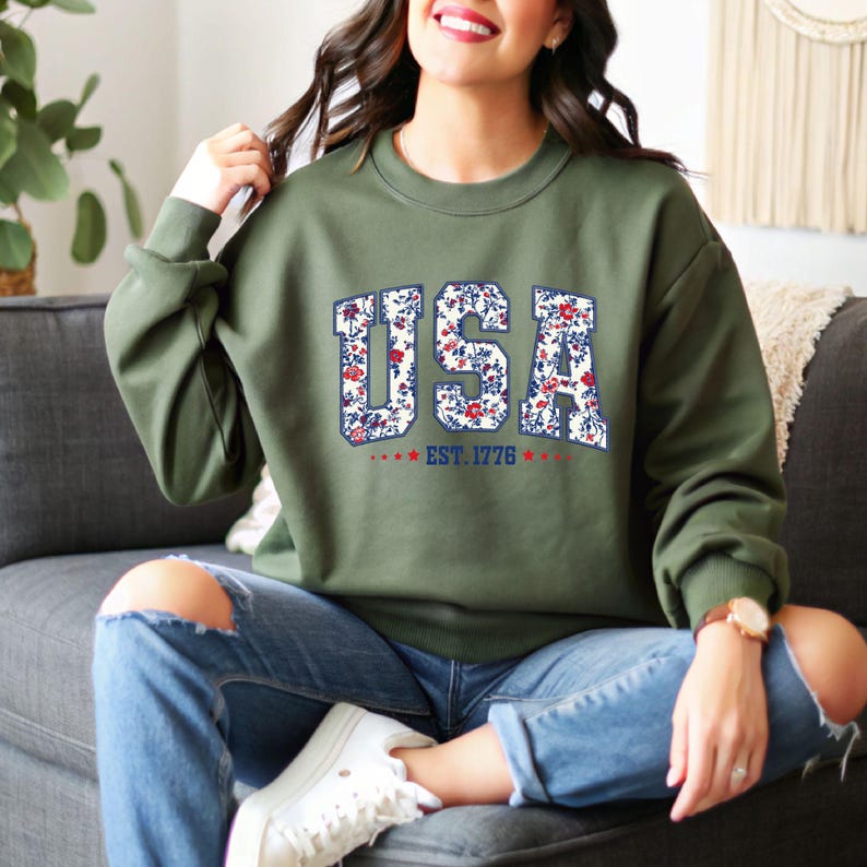May include: An olive green sweatshirt with the word "USA" in a floral pattern. The text "EST. 1776" is below the word "USA". The sweatshirt is being worn with blue jeans.