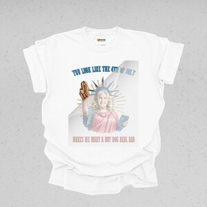 May include: White t-shirt with a graphic featuring a woman dressed as the Statue of Liberty holding a hot dog. Text reads "YOU LOOK LIKE THE 4TH OF JULY" and "MAKES ME WANT A HOT DOG REAL BAD."