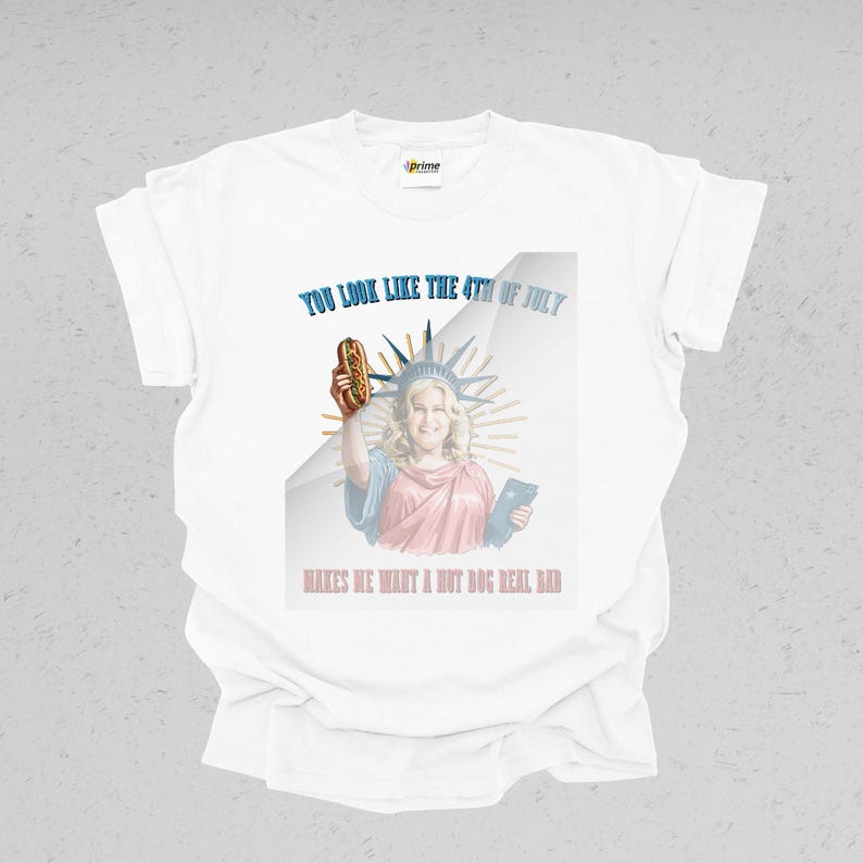 May include: White t-shirt with a graphic featuring a woman dressed as the Statue of Liberty holding a hot dog. Text reads "YOU LOOK LIKE THE 4TH OF JULY" and "MAKES ME WANT A HOT DOG REAL BAD."