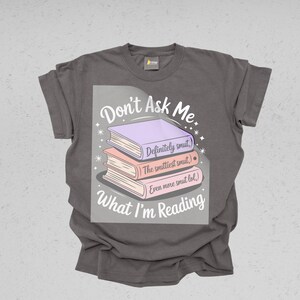 May include: A gray t-shirt with the text "Don't Ask Me What I'm Reading." The graphic features a stack of three books in purple, peach, and pink, with humorous text on each book. The shirt is a casual wear item.