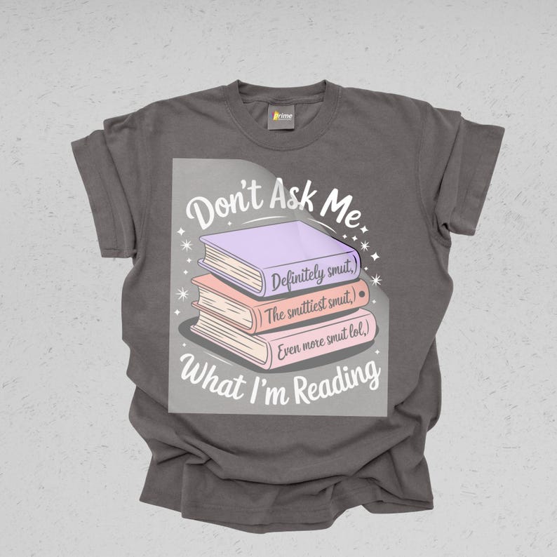 May include: A gray t-shirt with the text "Don't Ask Me What I'm Reading." The graphic features a stack of three books in purple, peach, and pink, with humorous text on each book. The shirt is a casual wear item.