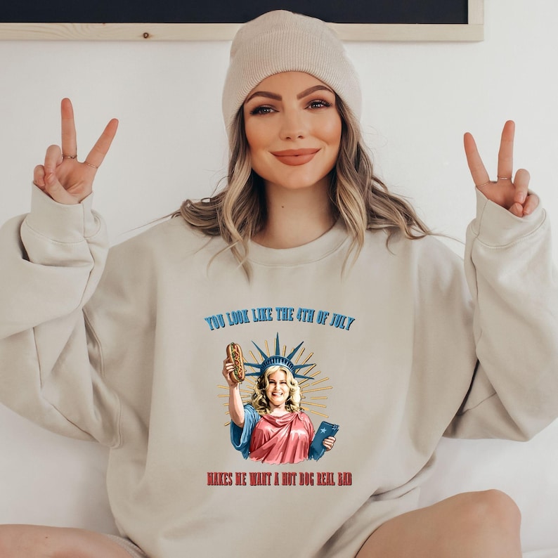 May include: Beige sweatshirt with the text "YOU LOOK LIKE THE 4TH OF JULY MAKES ME WANT A HOT DOG REAL BAD". The graphic features a cartoon of a woman holding a hot dog, styled as the Statue of Liberty. The person in the image is wearing a beige beanie.