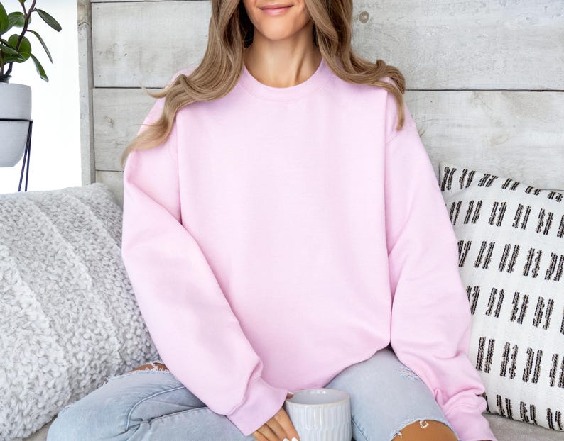 May include: A light pink crewneck sweatshirt. The sweatshirt is made of a soft material and has long sleeves. The person is wearing light blue jeans and holding a white mug. The background includes a white wooden wall and decorative pillows.