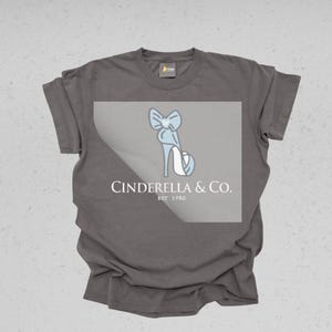 May include: A gray t-shirt featuring a light blue illustration of a high-heeled shoe with a bow. The text "Cinderella & Co. EST 1950" is printed below the shoe design in white.
