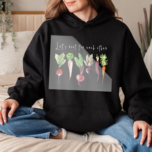 K&ouml;nnte beinhalten: Ein schwarzer Hoodie mit einer wei&szlig;en Grafik von Gem&uuml;se und dem Text "Let's root for each other".