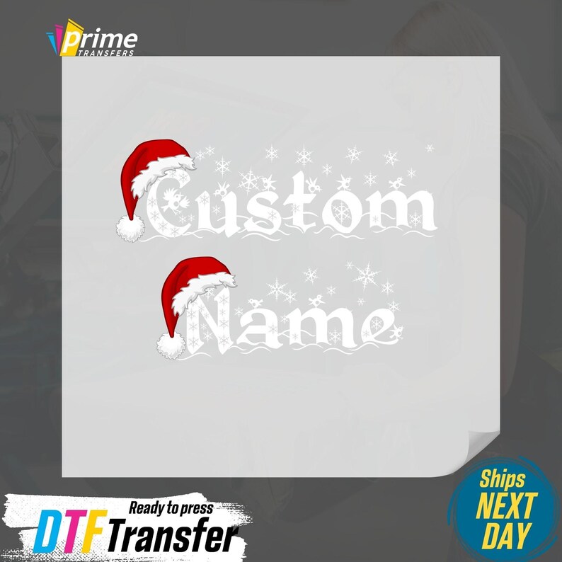 May include: A white graphic with the words "Custom Name" in a decorative font, each word topped with a Santa hat. Snowflakes are scattered around the text. The image is a ready-to-press DTF transfer.