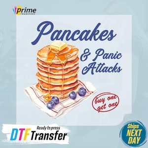 May include: A stack of pancakes with syrup and butter, surrounded by blueberries, on a red and white checkered napkin. The text "Pancakes & Panic Attacks" is above the pancakes, and "buy one get one" is in a red circle below. The text "Ready to press DTF Transfer" is below the napkin, and "Ships NEXT DAY" is in a blue circle on the right side of the image.