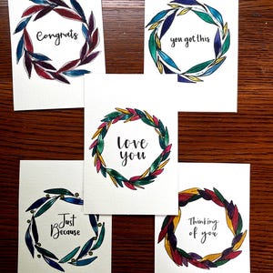 May include: Five white greeting cards with watercolor wreath designs. Each card has a different message: "Congrats," "you got this," "love you," "Just Because," and "Thinking of you." The wreaths feature colorful leaves in shades of blue, green, red, and yellow.
