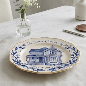 May include: A decorative plate featuring a blue and white house illustration, floral border, and gold trim. The plate is inscribed with "The Davies-Chen Family" and "Est 2008". The plate rests on a beige placemat.