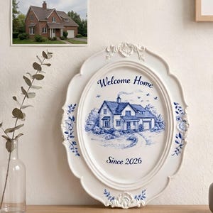 Könnte beinhalten: Weiße ovale Wandplakette mit einer blau-weißen Illustration eines Hauses, umrahmt von floralen Designs. Der Text "Welcome Home Since 2026" ist auf der Plakette zu sehen. Ein gerahmtes Foto eines Hauses ist ebenfalls sichtbar.