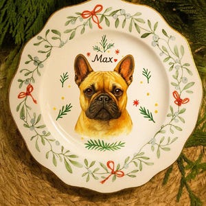 May include: A white ceramic plate with a decorative, scalloped edge. It features a watercolour portrait of a French Bulldog, surrounded by green foliage, red bows, and the name "Max".