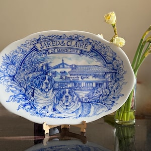 Pode incluir: Um prato oval de cerâmica branca com um design floral e arquitetônico azul. O prato apresenta o texto "JARED & CLAIRE 24 MARÇO 2026" e uma ilustração de um edifício e dois cães. O prato é exibido em um suporte de madeira.