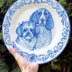 May include: A white ceramic plate featuring a blue ink illustration of two dogs, a Shih Tzu and a spaniel, centred on the surface. The rim of the plate is decorated with a detailed blue floral and scrollwork pattern that encircles the entire edge.