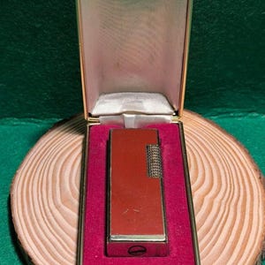 May include: A vintage, rectangular lighter in a gold-toned case with a red interior. The lighter is a reddish-brown color with a textured silver-colored ignition area. The case is open, revealing the lighter nestled in a red velvet lining.