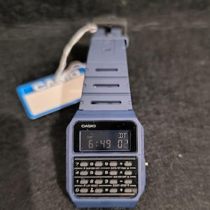 May include: A blue Casio calculator watch with a digital display showing the time. The watch has a calculator keypad below the screen and a blue strap. The Casio tag is attached to the watch.