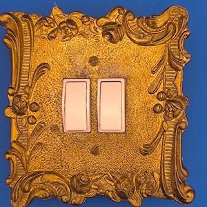 May include: A rectangular gold-colored light switch wall plate featuring an ornate, raised floral and scrollwork border. The center contains two white rocker-style light switches, secured by two visible screws positioned above and below the switches.