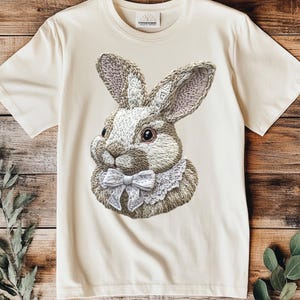 Op de afbeelding: Een crèmekleurig T-shirt met een geborduurde afbeelding van een bruin en wit konijn met een witte strik en een kanten kraag.