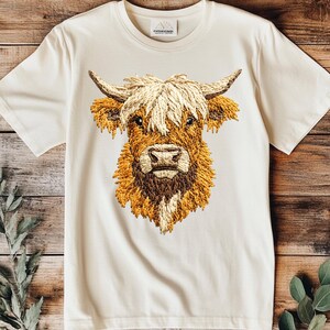 May include: A white t-shirt with an embroidered image of a Highland cow's head. The cow has a long, shaggy mane and a brown nose.