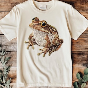 May include: A cream-colored t-shirt with a brown and green embroidered frog design.