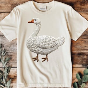 May include: A white t-shirt with a white goose embroidered on the front. The goose is facing the right side of the image. The t-shirt is on a wooden background.