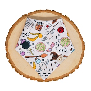 May include: White pet bandana with a repeating pattern of Harry Potter-themed illustrations, including owls, books, and a witch's hat. The text "Happee Birthdae Harry" is also featured. The bandana is displayed on a wooden slice.