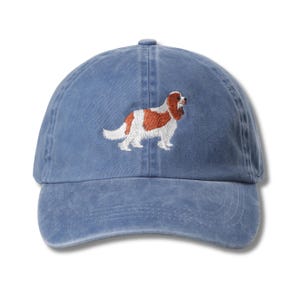 Könnte beinhalten: Eine blaue Baseballkappe mit gebogenem Schirm. Die Kappe hat einen gestickten Cavalier King Charles Spaniel Hund in Braun, Weiß und Beige. Der Hut ist aus einem weichen, gewaschenen Stoff gefertigt und hat einen verstellbaren Riemen.